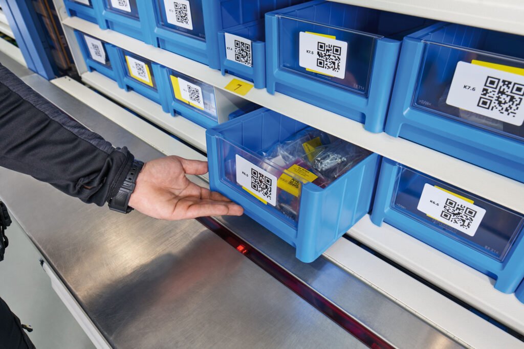 Empleado sacando un cajetín azul con piezas pequeñas en un sistema de almacenaje automatizado con etiquetas y códigos QR